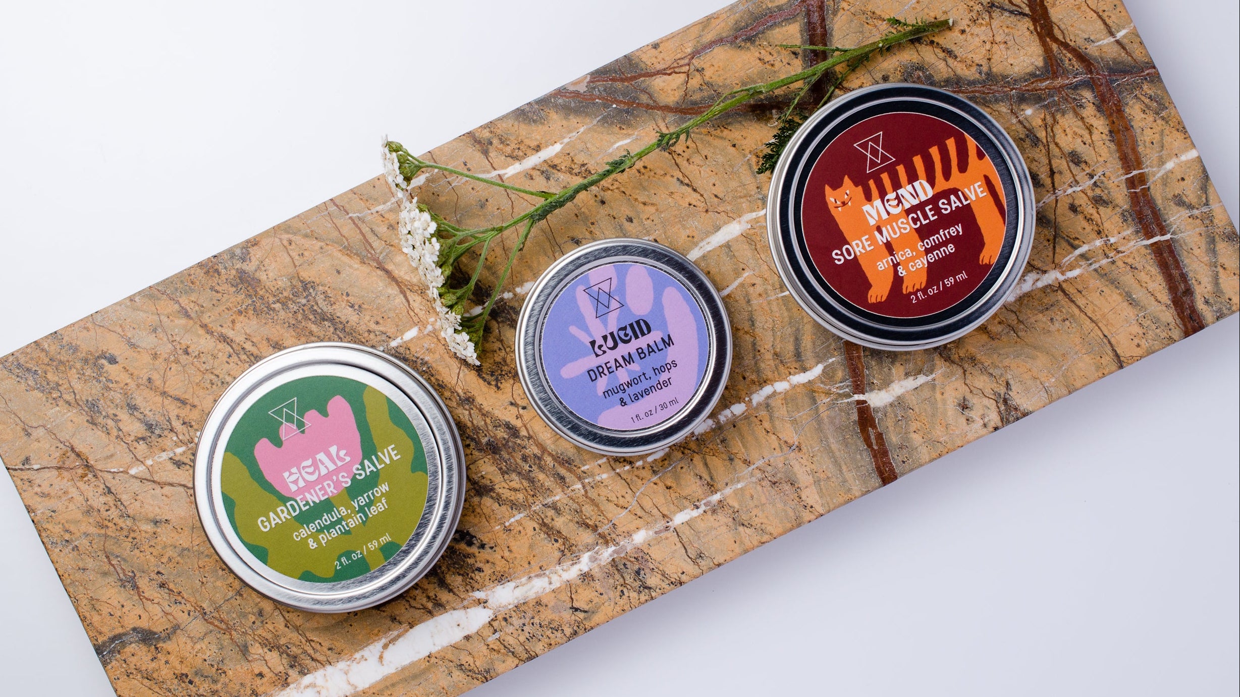 Three small round metal tins of balm and salve products arranged on a rectangular marble slab against a white background. Gardener's Salve, Dream Balm, and Sore Muscle Salve.
