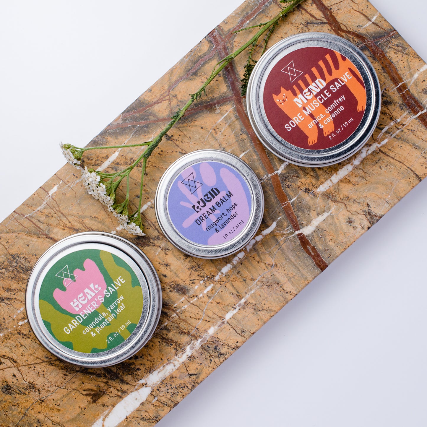 Three round metal tins of salves and balms on a marble slab: sore muscle salve, dream balm and gardener's salve. A sprig of yarrow lays beside them.