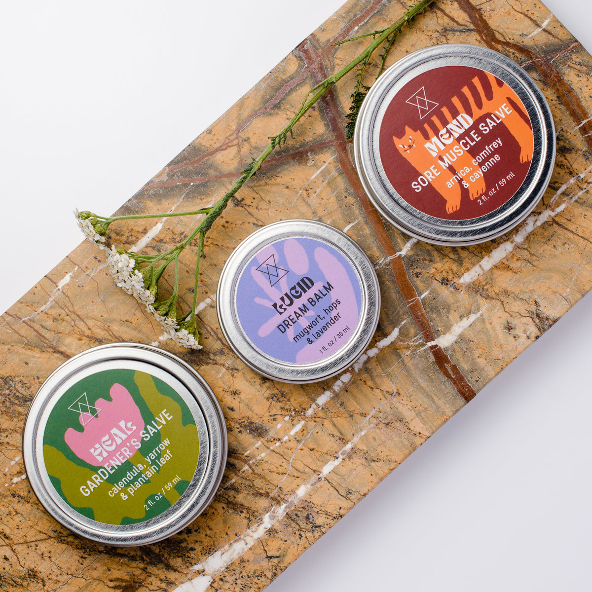 Three round metal tins of salves arranged on a brown marble board with a sprig of yarrow. The tins have colorful labels: green with pink, purple with blue, and orange with black.