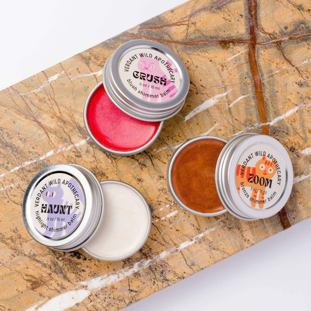 Three small round metal tins of shimmer balm... tins are open, showing pink, white, and bronze balms.