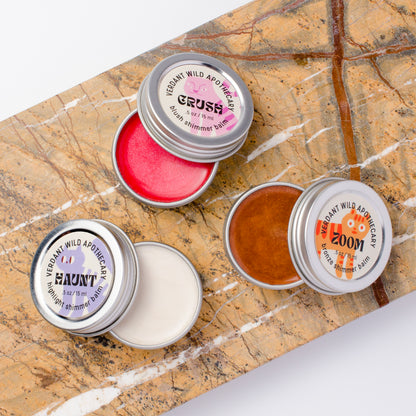 Three small round metal tins of shimmer balm... tins are open, showing pink, white, and bronze balms.