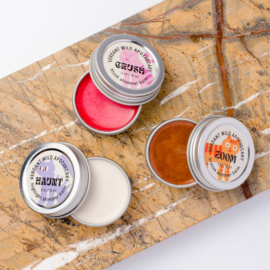 Three small round metal tins of shimmer balm... tins are open, showing pink, white, and bronze balms.