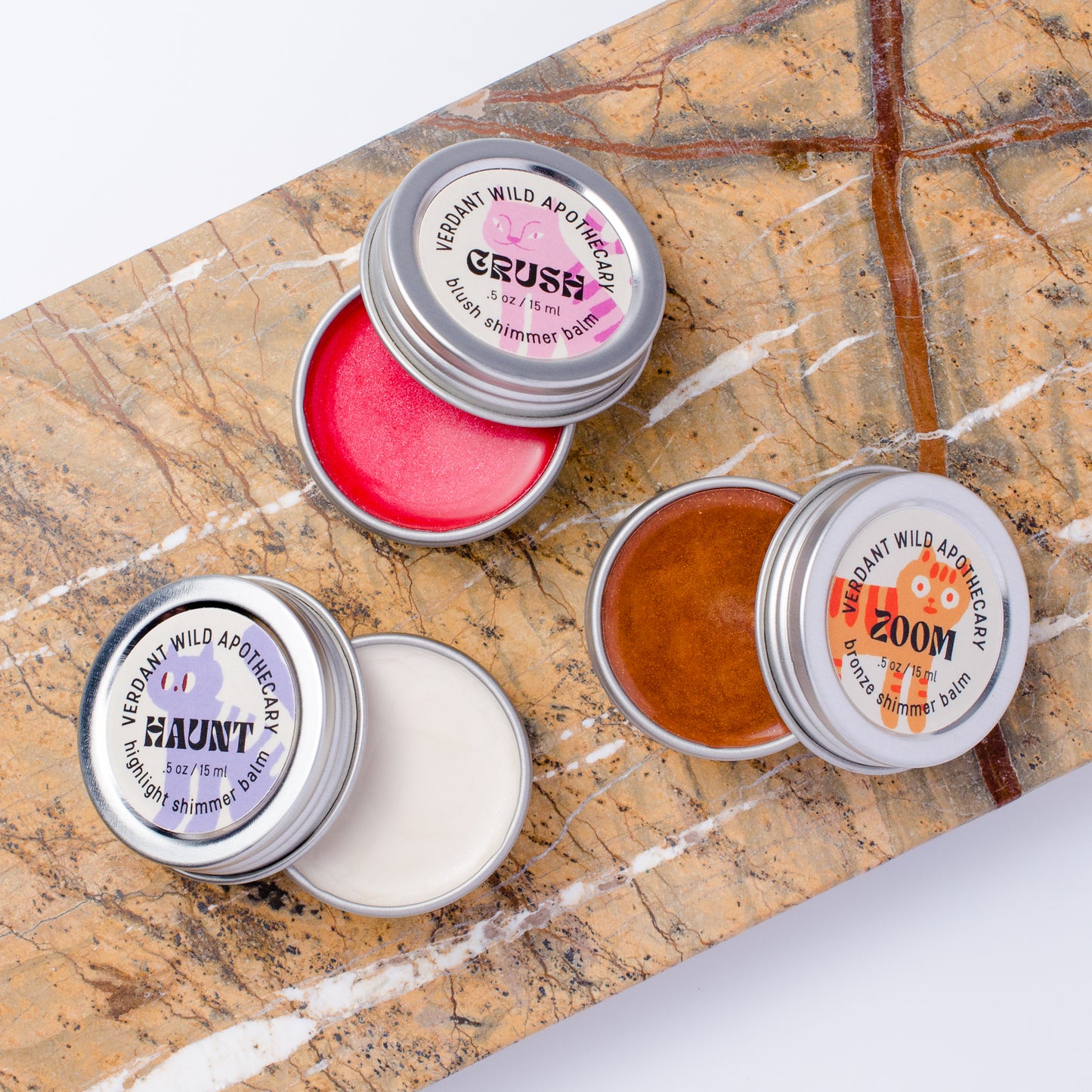 3 small round metal tin apothecary jars with silver lids are arranged on a brown marble slab. The tins are open, showing blush, highlighting and bronzing shimmer balms.