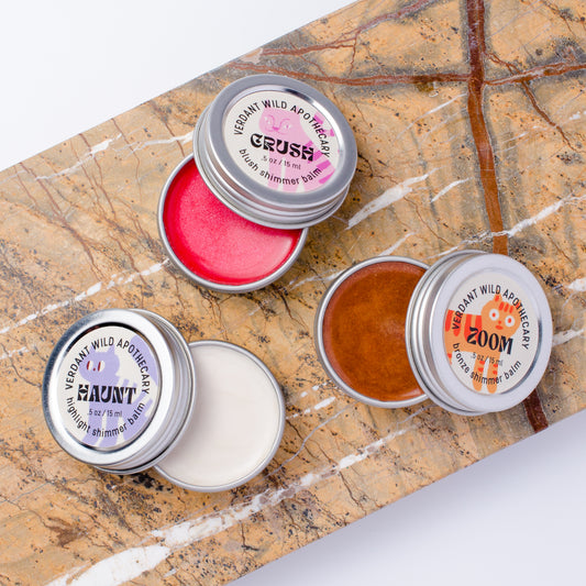 3 small round metal tin apothecary jars with silver lids are arranged on a brown marble slab. The tins are open, showing blush, highlighting and bronzing shimmer balms.