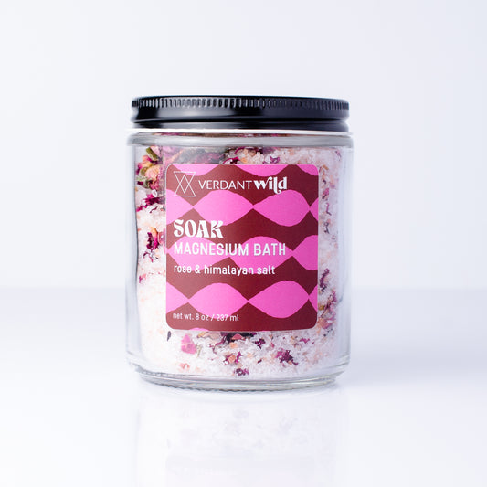 A glass jar of Verdant Wild Soak Magnesium Bath in rose & himalayan salt, featuring a black lid and a pink patterned label, set against a clean white background with a soft reflection.