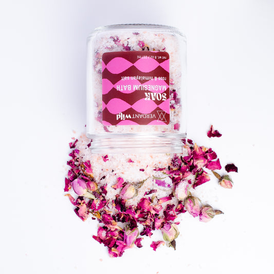 A clear glass jar labeled 'Soak Magnesium Bath' filled with pink bath salts and dried rose petals, spilled around the base on a white background.