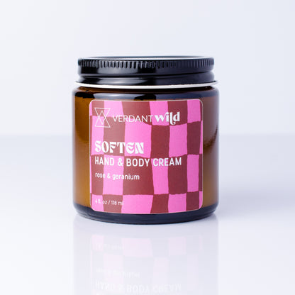 A jar of Verdant Wild Soften Hand & Body Cream with a pink and dark red checkered label, set against a clean white background with a soft reflection.