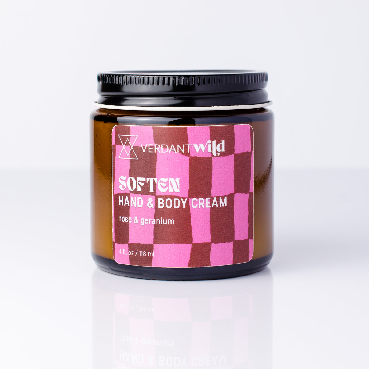 A jar of Verdant Wild Soften Hand & Body Cream with a pink and dark red checkered label, set against a clean white background with a soft reflection below.