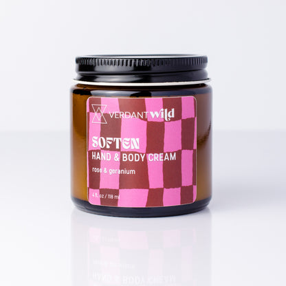 A jar of Verdant Wild Soften Hand & Body Cream with a pink and dark red checkered label, set against a clean white background with a soft reflection below.
