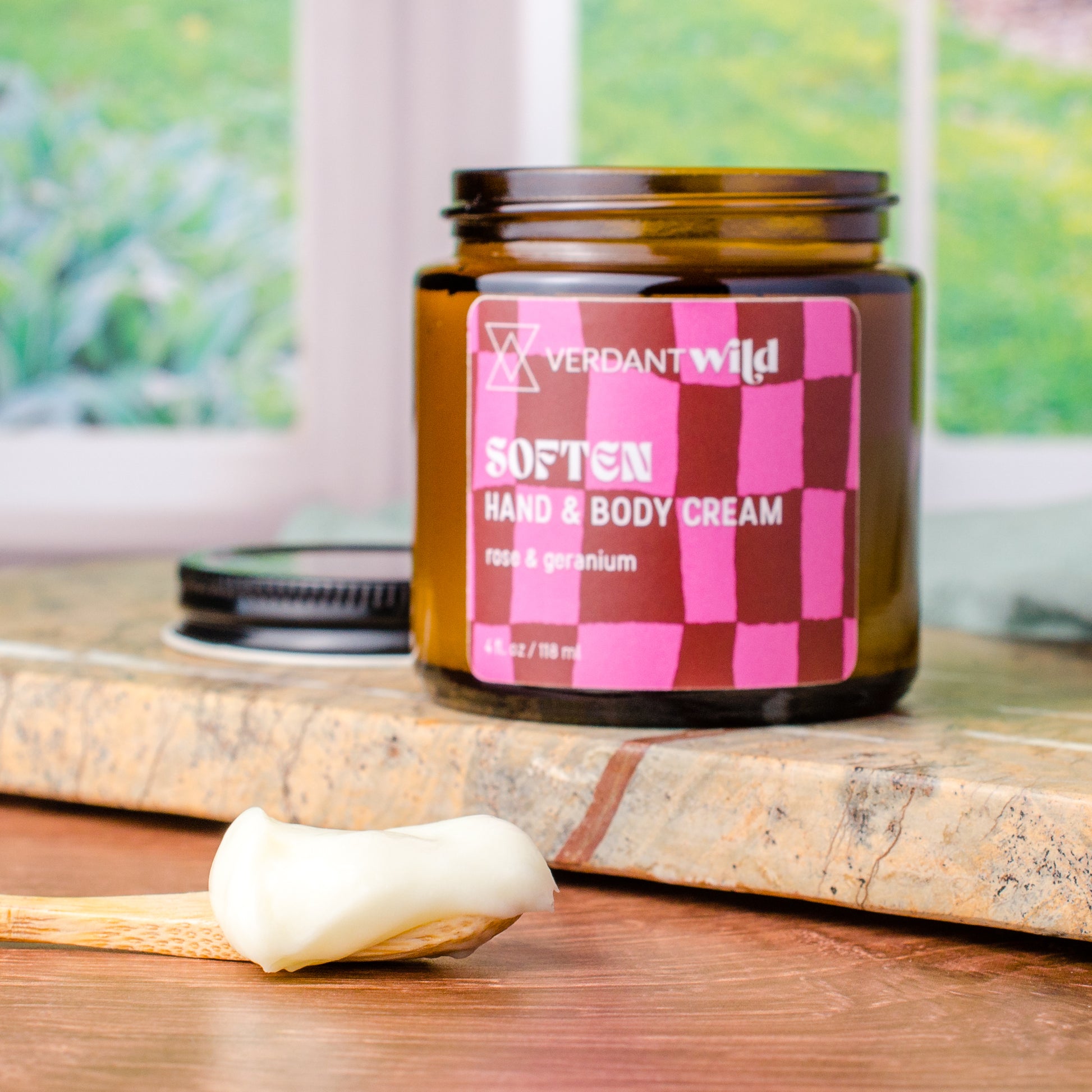A jar of Verdant Wild Soften Hand & Body Cream sits on a stone slab with a wooden spoon holding a dollop of the cream in the foreground. The jar is amber glass with a brown and pink checkered label.