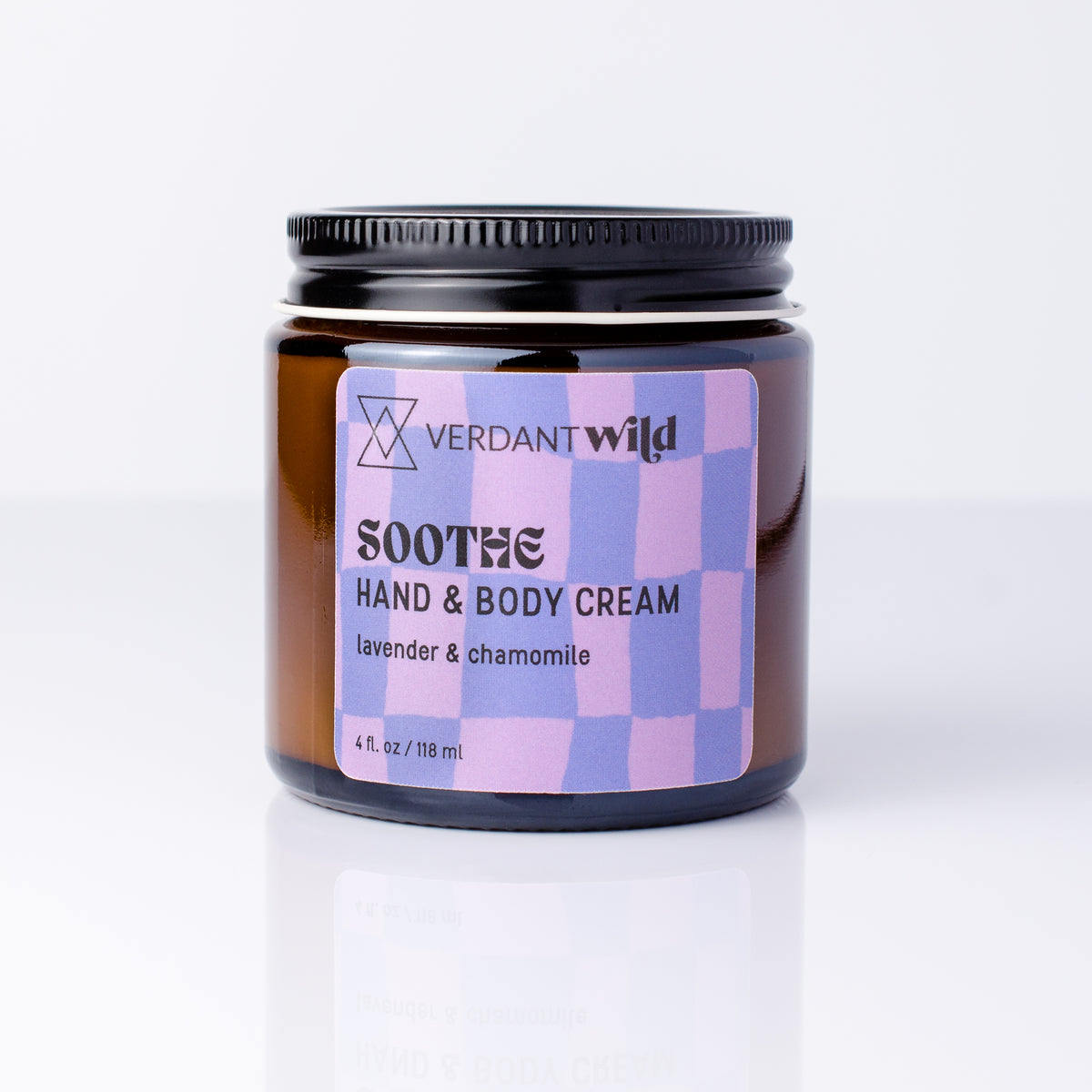 An amber glass jar of Verdant Wild Soothe Hand & Body Cream with a black lid and a lavender and purple checkered label, set against a clean white background with a soft reflection.