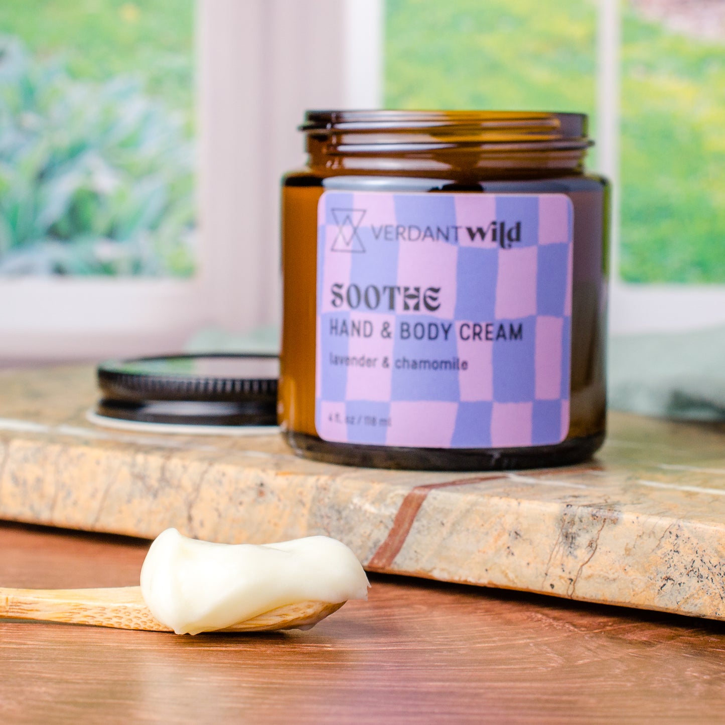 A jar of Verdant Wild 'Soothe' Hand & Body Cream sits on a stone slab with a wooden spoon holding a dollop of the white cream in the foreground. The background shows a softly blurred window and greenery.