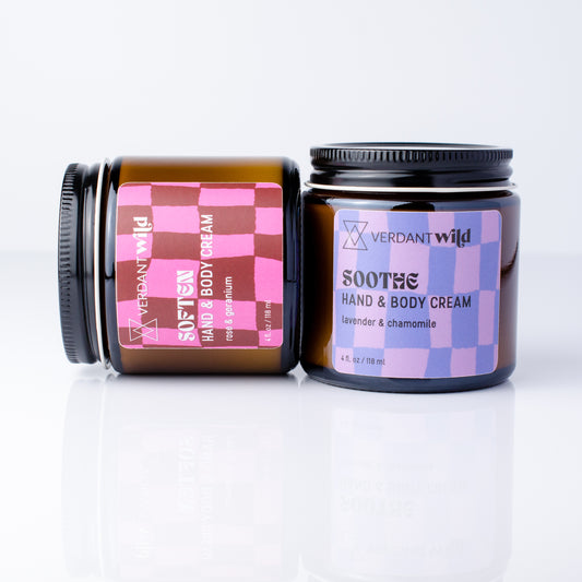 Two amber glass jars of Verdant Wild Hand & Body Cream on a white reflective surface. One jar is upright with a lavender and blue checkered label, and the other lies on its side showing the same label design in dark red and bright pink.