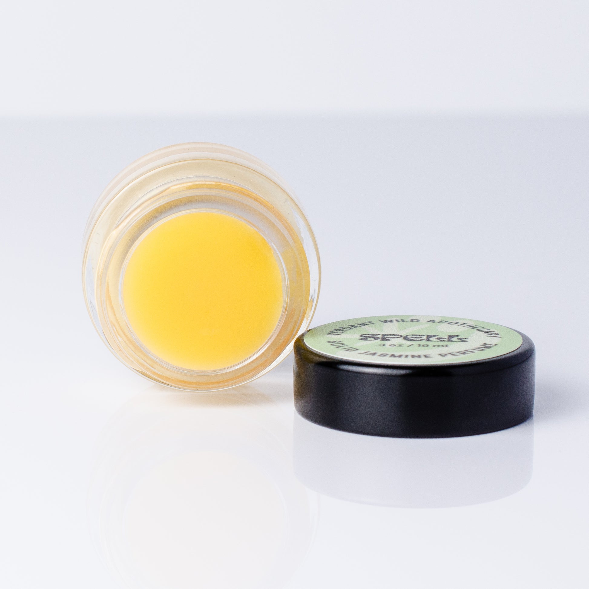 A small round jar of solid jasmine perfume shown open with the lid beside it on a clean white background. 