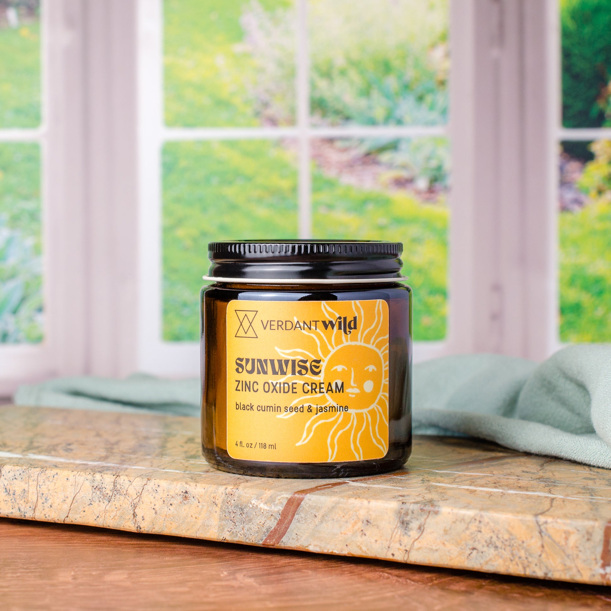 A jar of Verdant Wild Sunwise Zinc Oxide Cream sits on a stone countertop with a blurred kitchen window and greenery in the background.