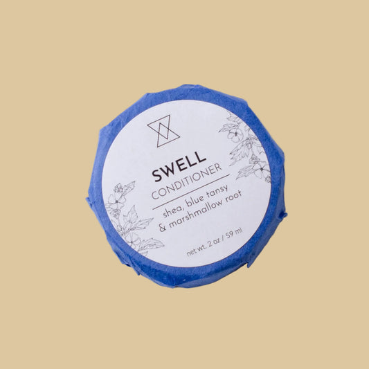 A round wrapped bar of Swell Conditioner soap on a clean background. The bar is wrapped in blue paper and features a white circular label with black text reading 'Swell Conditioner' and botanical line drawings.