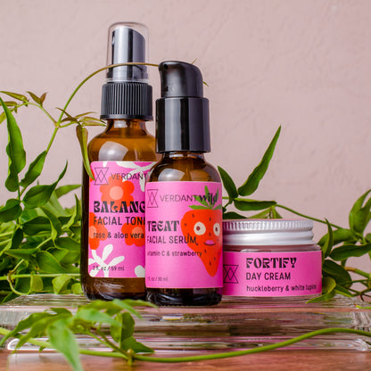 Three skincare products from Verdant Beauty arranged on a glass tray with green leaves: a facial toner bottle, a facial serum bottle, and a day cream jar, all with bright pink labels against a soft blush background.