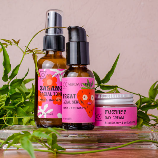 Three skincare products from Verdant Beauty arranged on a glass tray with green leaves: a facial toner bottle, a facial serum bottle, and a day cream jar, all with bright pink labels against a soft blush background.