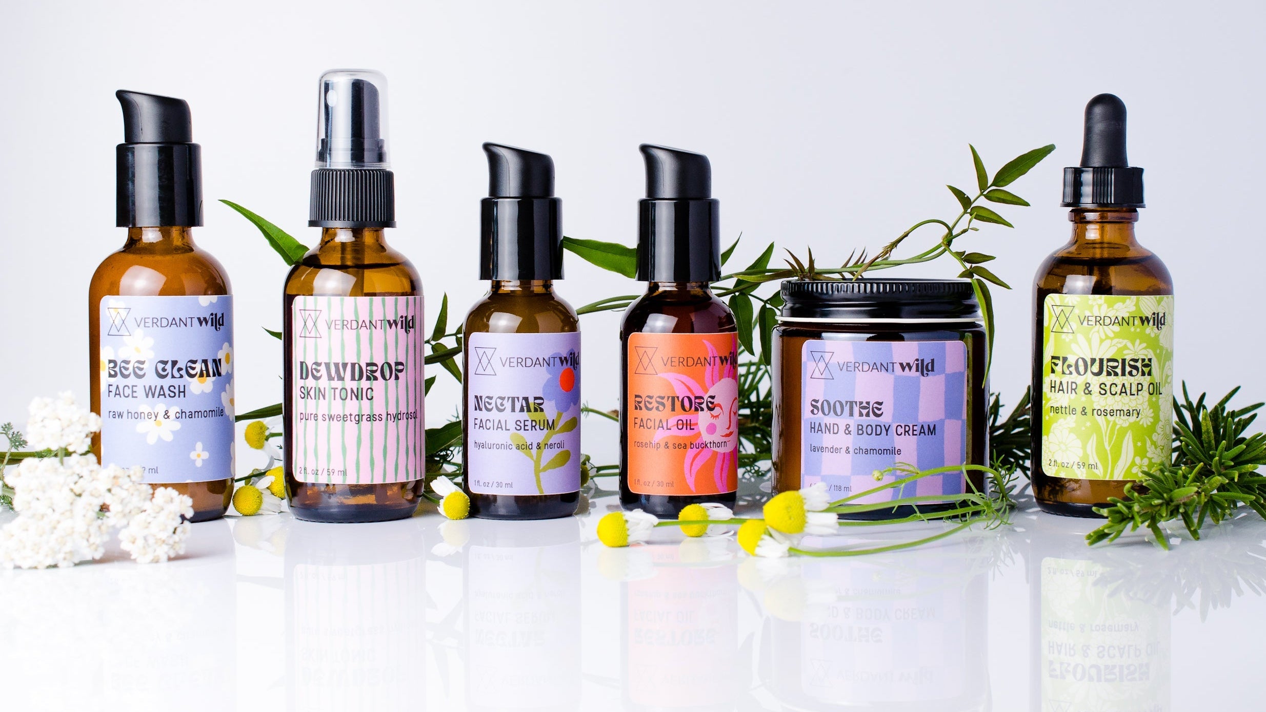 Six skincare products from the brand Verdant Wild are lined up on a reflective white surface against a light gray background, styled with green leaves and small yellow flowers. The products include face wash, body wash, facial toner, facial cleanser, hand & body cream, and hair & scalp oil in amber glass bottles and a jar with colorful labels.