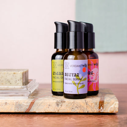 Three small cosmetic bottles with black pumps are arranged on a stone slab beside a bar of soap. The center bottle is labeled 'VERDANT WILD NECTAR FACIAL SERUM,' with two additional bottles behind it partially visible.