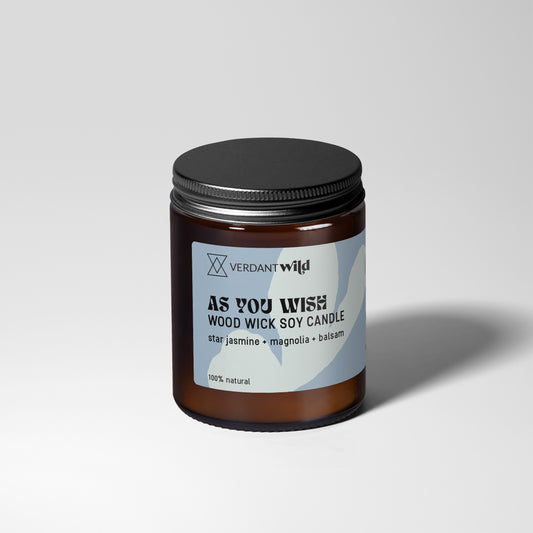 A Verdant Wild 'As You Wish' wood Wick soy candle in an amber glass jar with a black lid and a light blue label, set against a neutral gray background.