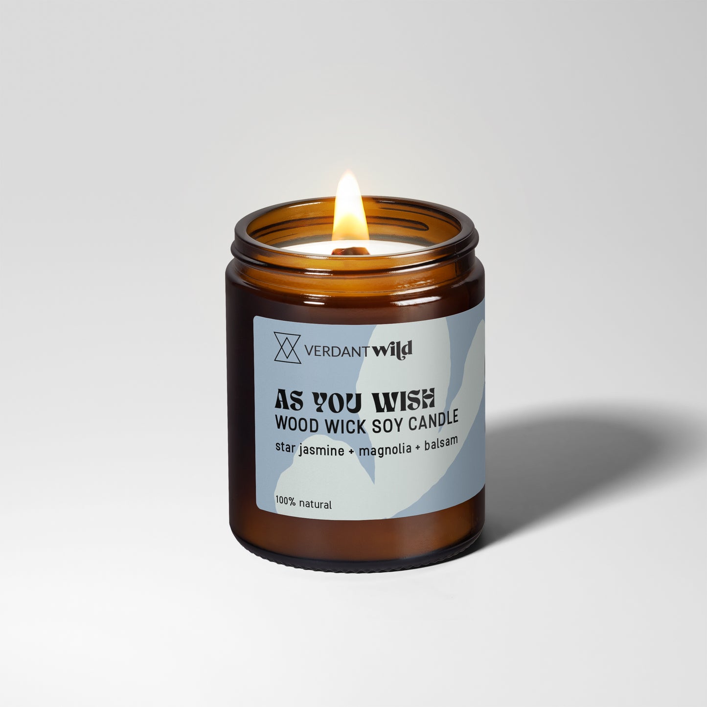A lit amber glass jar candle with a white and blue label reading 'VERDANT WILD AS YOU WISH WOOD WICK SOY CANDLE' on a light gray studio background.