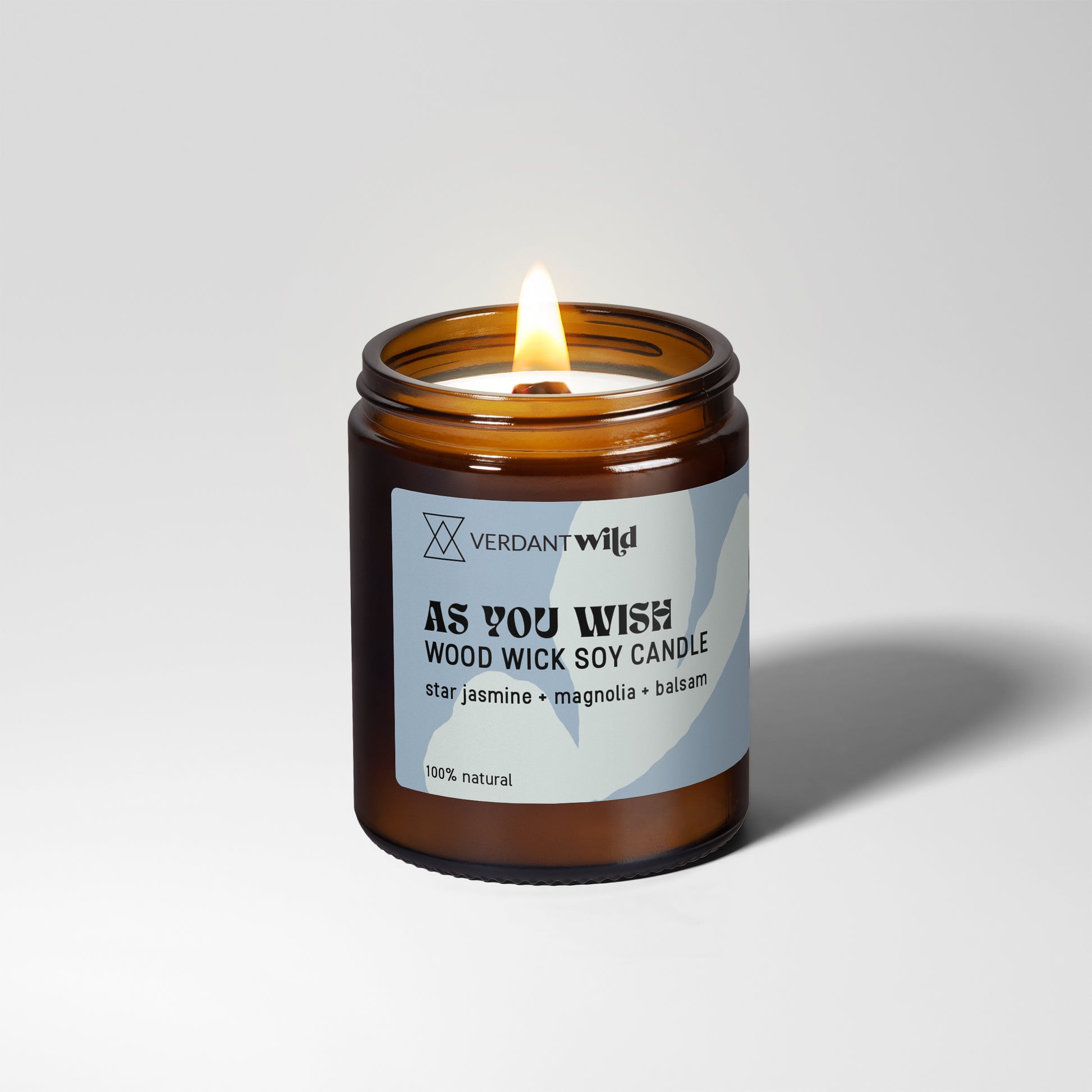 A lit amber glass jar candle with a white and blue label reading 'VERDANT WILD AS YOU WISH WOOD WICK SOY CANDLE' on a light gray studio background.