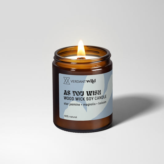 A lit amber glass jar candle with a white and blue label reading 'VERDANT WILD AS YOU WISH WOOD WICK SOY CANDLE' on a light gray studio background.