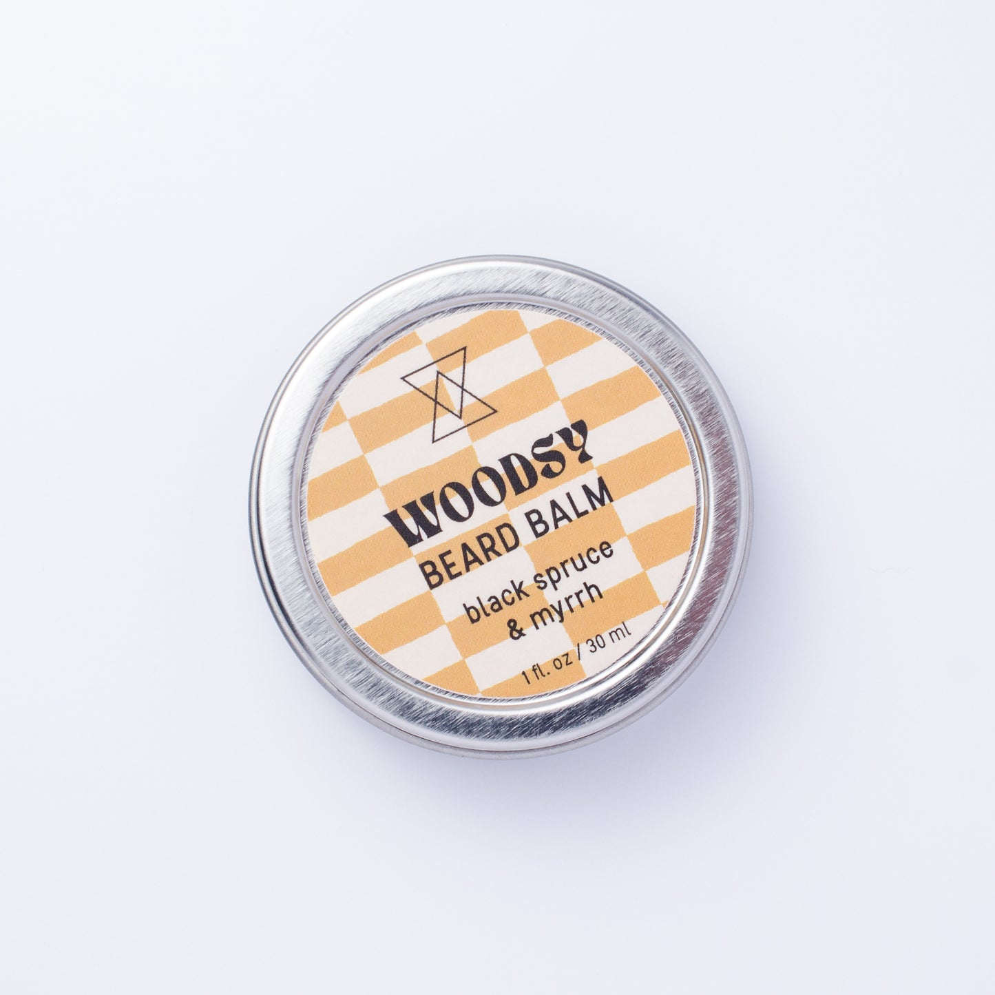 A round silver tin of WOODSY Beard Balm in the 'black spruce & myrrh' scent, featuring a brown and white checkered label, photographed from directly above on a clean white background.