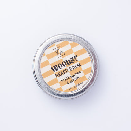 A round silver tin of WOODSY Beard Balm in the 'black spruce & myrrh' scent, featuring a brown and white checkered label, photographed from directly above on a clean white background.