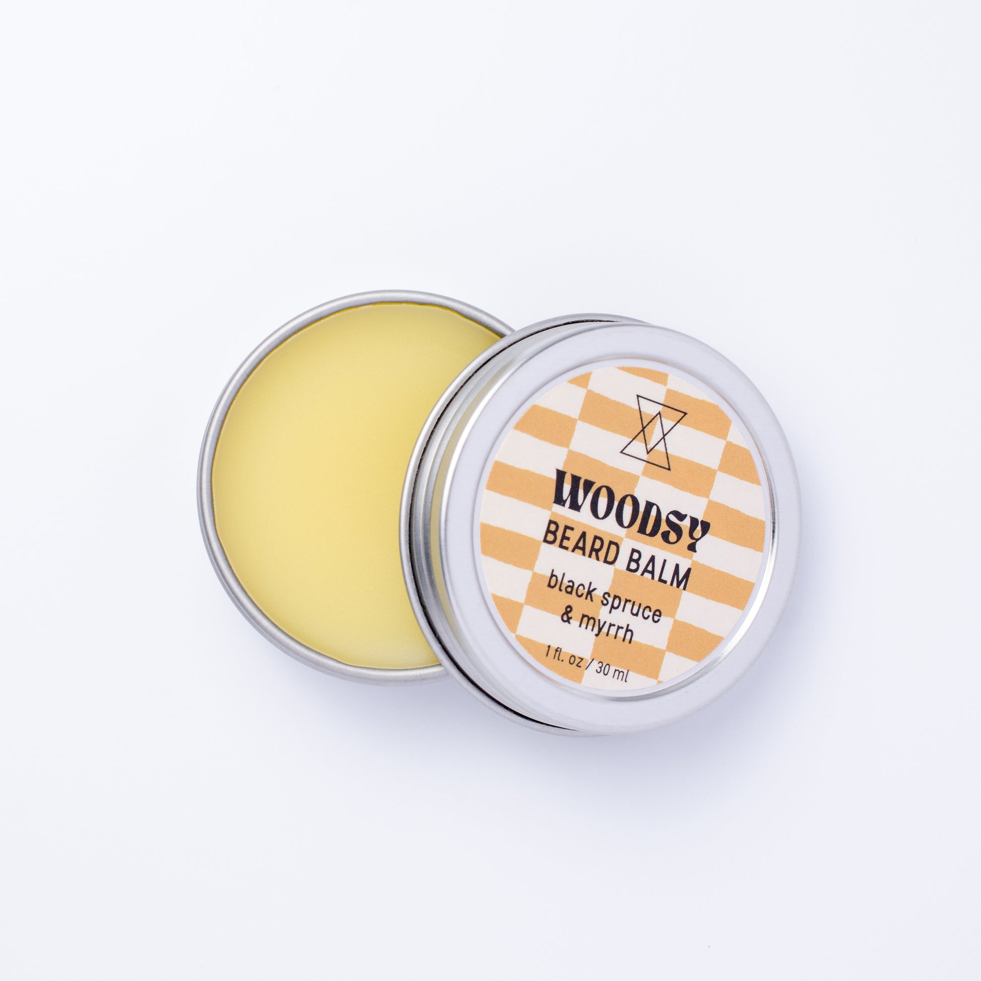 An open round metal tin of Woodsy Beard Balm showing the pale yellow balm inside, with the lid beside it featuring a white and brown checkered label.