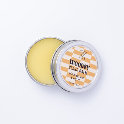 An open round metal tin of Woodsy Beard Balm showing the pale yellow balm inside, with the lid beside it featuring a white and brown checkered label.