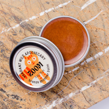 An open round metal tin of shimmer balm lies on a beige-and-brown marble surface, with the lid placed beside it. The label reads 'bronze shimmer balm.