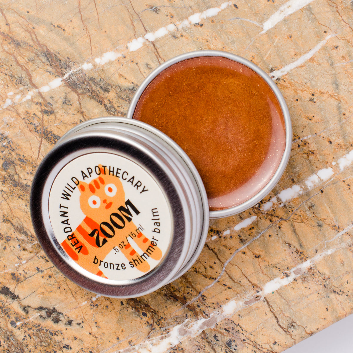 An open round metal tin of shimmer balm lies on a beige marble surface, with the lid beside it. The label reads 'Zoom bronze shimmer balm' with an orange cat illustration.